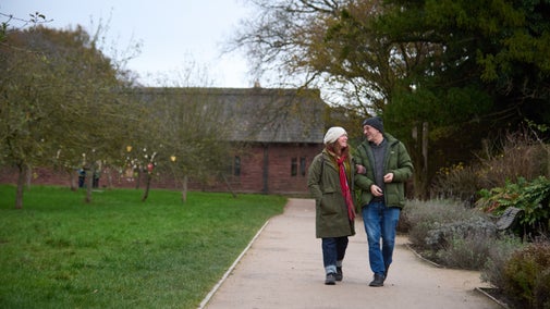 Two people walking along a wide gravel path in a park, wearing winter coats and hats. There are bare trees and green grass on either side, with a red-brick building in the background.